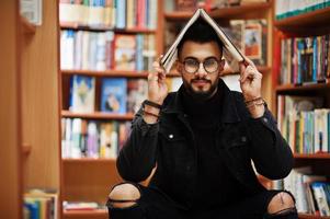 grand étudiant arabe intelligent, portant une veste en jean noir et des lunettes, assis à la bibliothèque et se couvrant la tête avec un livre. photo