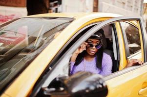 femme afro-américaine en robe violette et casquette posée sur une voiture jaune. photo