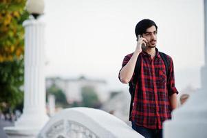 jeune étudiant indien en chemise à carreaux et jeans avec sac à dos parlant sur téléphone portable. photo