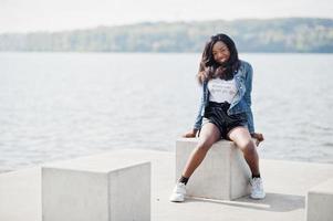 modèle mince afro-américain à la peau foncée posé dans un short noir et une veste en jean contre le bord de mer. photo