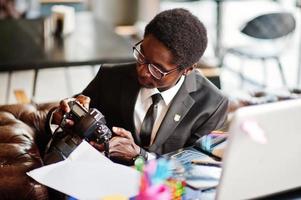 photographe afro-américain paparazzi homme portant un costume noir et des lunettes assis au bureau avec appareil photo et travaillant derrière un ordinateur portable.