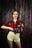 jeune femme au foyer drôle en chemise à carreaux et short jaune pin up style avec casserole sur fond en bois. photo