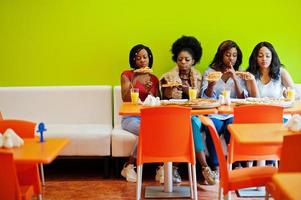 quatre jeunes filles africaines dans un restaurant aux couleurs vives mangeant des tranches de pizza dans les mains. photo