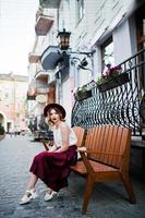 fille modèle blonde à la mode et belle en élégante jupe en velours rouge, chemisier blanc et chapeau, posée sur un banc dans la rue de la ville. photo