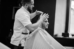 jeune homme barbu se coupe les cheveux par un coiffeur assis sur une chaise au salon de coiffure. âme de barbier. photo