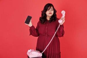 jolie femme sud-asiatique en robe rouge foncé posée au studio sur fond rose et utilise un vieux téléphone vintage avec un téléphone mobile moderne. photo