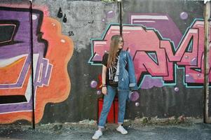 élégante fille hipster décontractée en jeans et lunettes contre un grand mur de graffitis. photo