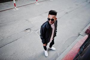 homme barbe indien élégant à la veste en cuir noir et lunettes de soleil. modèle indien posé en plein air dans les rues de la ville. photo