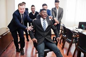 six hommes d'affaires multiraciaux debout au bureau et roulent l'homme sur une chaise. groupe diversifié d'employés masculins en tenue de soirée s'amusant. photo