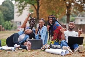 groupe de cinq étudiants africains qui passent du temps ensemble sur le campus de la cour universitaire. amis afro noirs assis sur l'herbe et étudiant avec des ordinateurs portables. photo