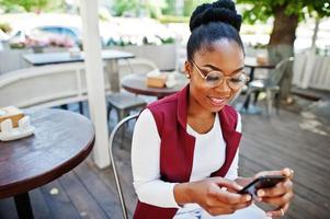 une fille afro-américaine porte des lunettes avec un téléphone portable assis au café en plein air.
