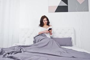 portrait en studio d'une jeune fille brune en blouse blanche allongée sur le lit et utilisant un téléphone portable. photo