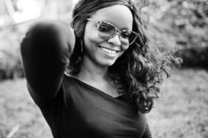 portrait en gros plan d'une fille afro-américaine à lunettes de soleil, vêtements noirs posés à l'extérieur. femme noire à la mode. photo