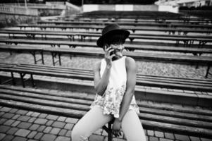 incroyable femme modèle afro-américaine en pantalon vert et chapeau noir posé au banc et à l'aide d'un smartphone. photo