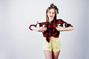 jeune femme au foyer drôle en chemise à carreaux et short jaune style pin up avec deux casseroles isolées sur fond blanc. photo