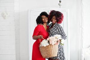 deux femmes afro-américaines à la mode en robe de soirée debout avec un panier de fleurs sur les mains contre une vieille armoire vintage à la salle blanche. photo