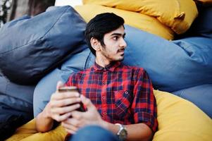 jeune étudiant indien en chemise à carreaux et jeans assis et se détendre sur des oreillers extérieurs. passer du temps avec le téléphone portable. photo