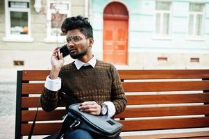 portrait d'un modèle de jeune homme indien élégant pose dans la rue, assis sur un banc avec sac à main et smartphone. photo