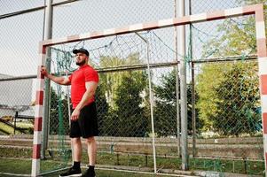 jeune homme musclé barbu brutal portant une chemise rouge, un short et une casquette au stade. photo