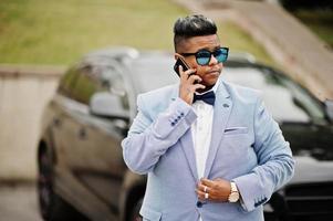 homme arabe élégant en veste, noeud papillon et lunettes de soleil contre une voiture suv noire. riche homme d'affaires arabe parlant sur téléphone mobile. photo