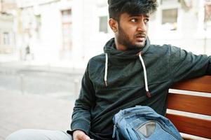 portrait d'un jeune mannequin indien élégant pose dans la rue, assis sur un banc et tient le smartphone à portée de main. photo