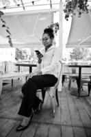 une fille afro-américaine porte des lunettes avec un téléphone portable assis au café en plein air. photo