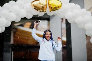 élégante fille afro-américaine avec des dreads tenant un téléphone portable à portée de main, en plein air contre une arche de ballons par temps de neige. photo