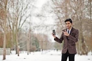 homme étudiant indien élégant en costume marron et lunettes posées à la journée d'hiver en plein air avec un téléphone portable à portée de main. photo