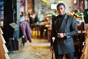 élégant gentleman afro-américain dans une élégante veste noire, tenant un bâton de marche rétro comme flacon de canne ou canne basculante avec poignée en boule de diamant doré. riche homme afro à la mode. photo