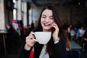 fille brune assise sur un café avec une tasse de cappuccino et parlant de téléphone portable. photo