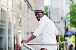 homme afro-américain élégant en chemise blanche et pantalon coloré avec chapeau et lunettes posés à l'extérieur. garçon modèle à la mode noir. photo