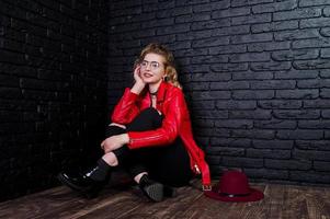 portrait en studio d'une fille blonde avec un chapeau rouge, des lunettes et une veste en cuir posées contre un mur de briques. photo