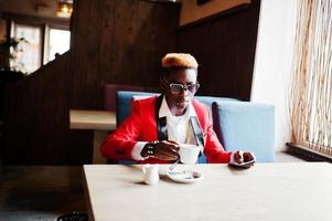 modèle d'homme afro-américain de mode au costume rouge, avec des cheveux en surbrillance et des lunettes de soleil assis au café avec du café. photo