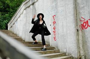 fille sensuelle tout en noir, lèvres rouges et chapeau. une femme dramatique gothique tient une fleur de chrysanthème blanche contre un mur de graffitis. photo