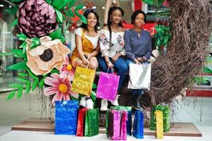 belles trois filles afro-américaines bien habillées avec des sacs à provisions colorés assis sur la zone photo de décoration de printemps dans le centre commercial.