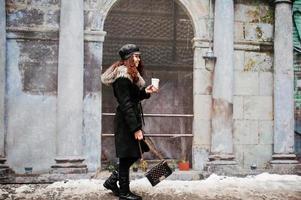 fille mexicaine bouclée en casquette en cuir et tasse de café en plastique à portée de main marchant dans les rues de la ville. photo