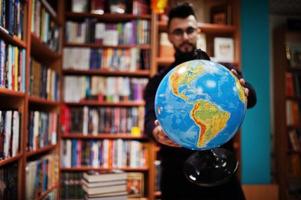 grand étudiant arabe intelligent, portant un col roulé violet et des lunettes, à la bibliothèque tenant un globe terrestre à portée de main. photo