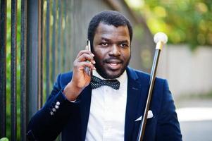 bel homme afro-américain à la mode en tenue de soirée et noeud papillon avec canne parlant au téléphone. photo