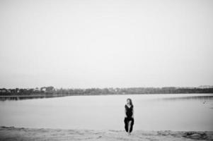 femme aux pieds nus sensuelle blonde en robe de marsala rouge posant contre le lac sur le sable. photo