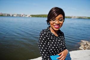 élégante fille modèle afro-américaine à lunettes, jupe bleue et chemisier noir posé en plein air sur le pont contre le lac. photo