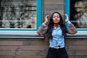 fille afro-américaine hipster portant une chemise en jean à manches léopard posant dans la rue contre une maison en bois avec des fenêtres. photo