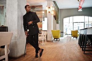 élégant gentleman afro-américain dans une élégante veste noire, tenant un bâton de marche rétro comme flacon de canne ou canne basculante avec poignée en boule de diamant doré. riche homme afro à la mode. photo