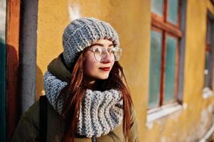 portrait de jeune fille brune en écharpe et chapeau gris, lunettes par temps froid avec soleil contre le mur orange de la vieille maison. photo