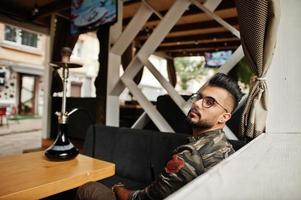 homme arabe à barbe élégante dans des verres et une veste militaire fumant le narguilé au bar de la rue. modèle arabe se reposant. photo