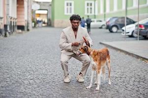 homme afro élégant en costume beige old school avec chien barzoï russe. jeune homme africain à la mode en veste décontractée sur torse nu. photo