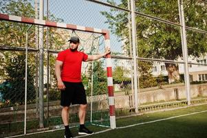 jeune homme musclé barbu brutal portant une chemise rouge, un short et une casquette au stade. photo