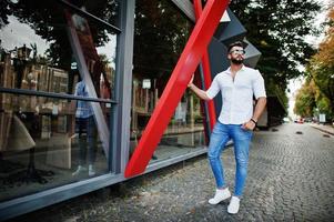 grand modèle d'homme arabe élégant en chemise blanche, jeans et lunettes de soleil posés dans la rue de la ville. Barbe mec arabe attrayant. photo