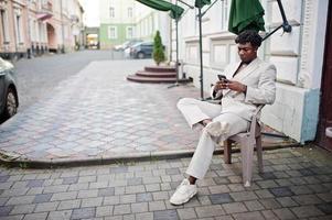 homme afro élégant en costume beige old school assis en plein air sur une chaise avec un téléphone portable à portée de main. jeune homme africain à la mode en veste décontractée sur torse nu. photo