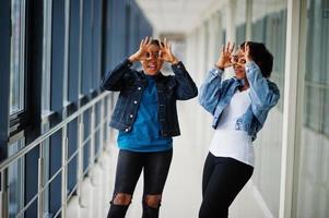 deux amies africaines en veste de jeans montrant des lunettes par les doigts à l'intérieur ensemble. photo