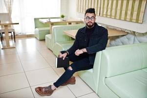 un homme arabe porte une veste en jean noir et des lunettes assis dans un café, lit un livre. mec modèle arabe élégant et à la mode. photo